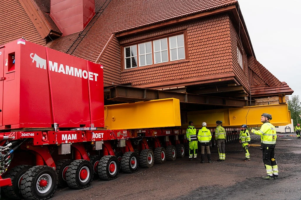 113-letni kościół w Szwecji zmieni swoje miejsce. Przewiozą całą budowlę na platformach Przesuwanie dużego budynku za pomocą zaawansowanego, wieloosiowego pojazdu z czerwonym modułem firmy Mammoet oraz żółtymi belkami wsporczymi, kilku pracowników w żółtych ubraniach ochronnych nadzoruje operację.