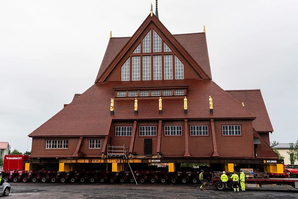 Wielkie przedsięwzięcie w Szwecji. Transport kościoła ma zająć dwa dni Duży drewniany budynek kościoła umieszczony na specjalnych platformach transportowych, podparty metalowymi konstrukcjami i otoczony przez kilku pracowników w odblaskowej odzieży, przygotowujących obiekt do przemieszczenia.