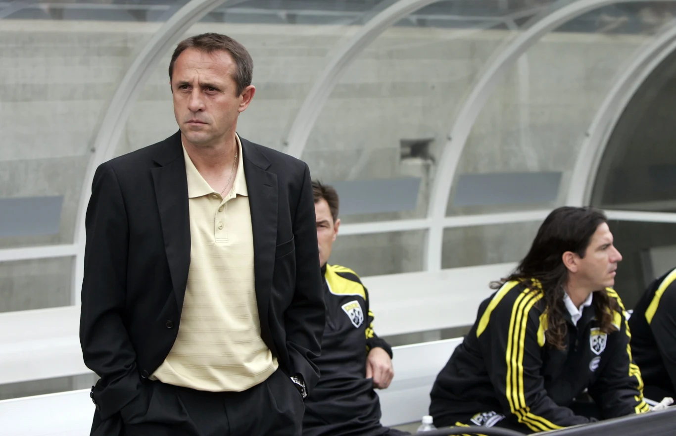 Robert Warzycha zapisał się w historii jako strzelec pierwszego "polskiego" gola w Premier League Trener w ciemnej marynarce i kremowej koszuli stoi przy ławce rezerwowych na stadionie piłkarskim, w tle kilku piłkarzy w klubowych dresach siedzi na ławce, wokół przezroczyste zadaszenie.