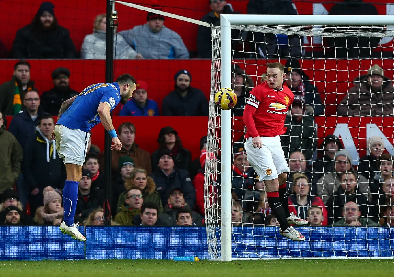 Ponad 22 lata kibice czekali na kolejnego gola strzelonego przez Polaka w Premier League. Impas przerwał Marcin Wasilewski w meczu z Manchesterem United Piłkarz w niebieskim stroju wyskakuje do główki w pobliżu bramki, podczas gdy zawodnik w czerwonej koszulce Manchesteru United i bramkarz obserwują lot piłki przed bramką. W tle widoczna trybuna z kibicami.