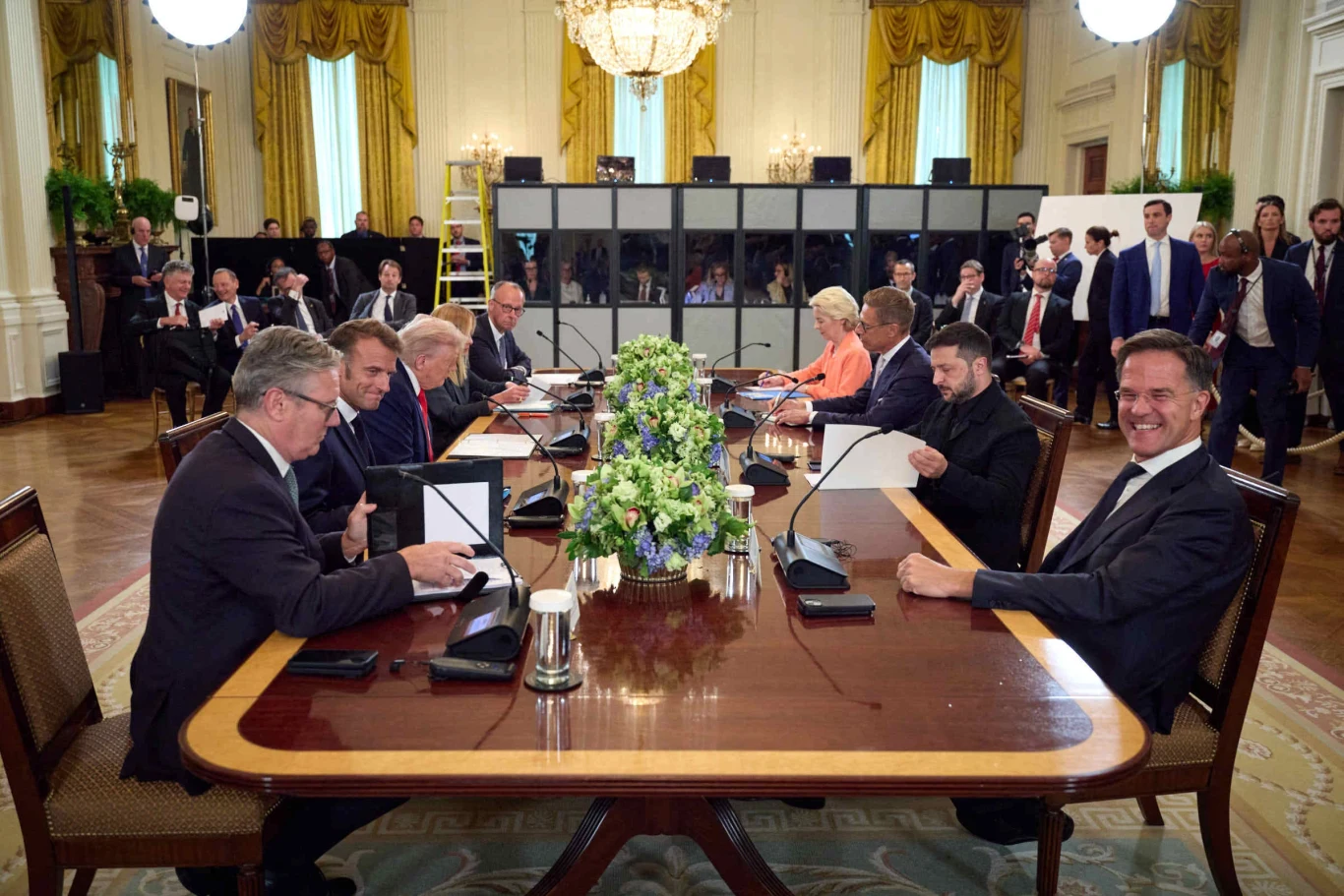 Poniedziałkowe spotkanie prezydenta USA Donalda Trumpa z przywódcami europejskimi Grupa polityków i urzędników siedzących wokół dużego stołu konferencyjnego, w centrum stołu duża kompozycja kwiatowa, wszyscy skupieni na podpisywaniu dokumentów lub rozmowie, w tle obecni są obserwatorzy i dziennikarze.