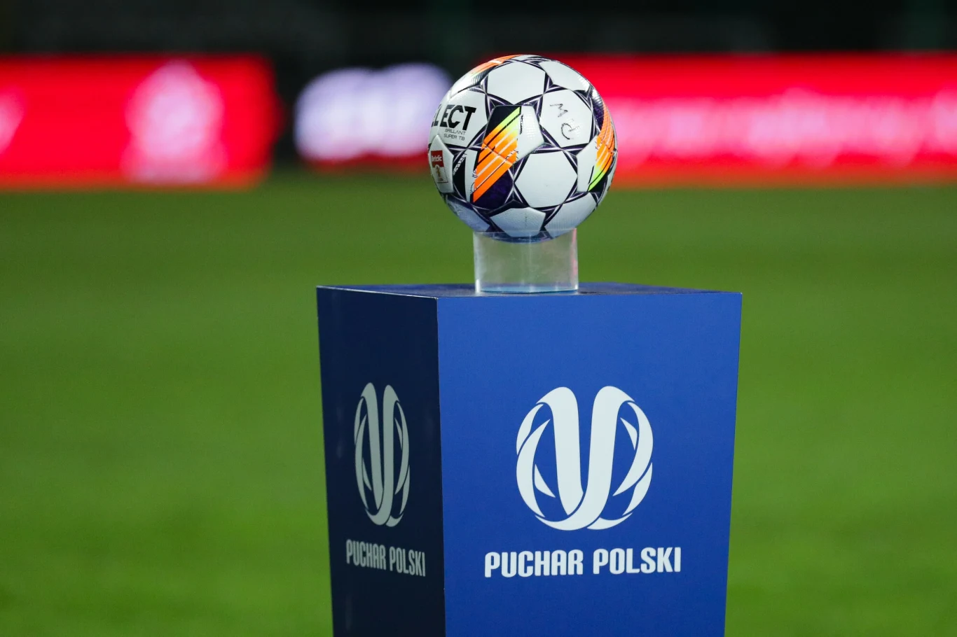 Oficjalna piłka meczowa na podstawie ekspozycyjnej, ustawiona na granatowym cokole z białym logo i napisem "Puchar Polski", rozmyte tło stadionu z czerwonymi elementami podświetlenia.