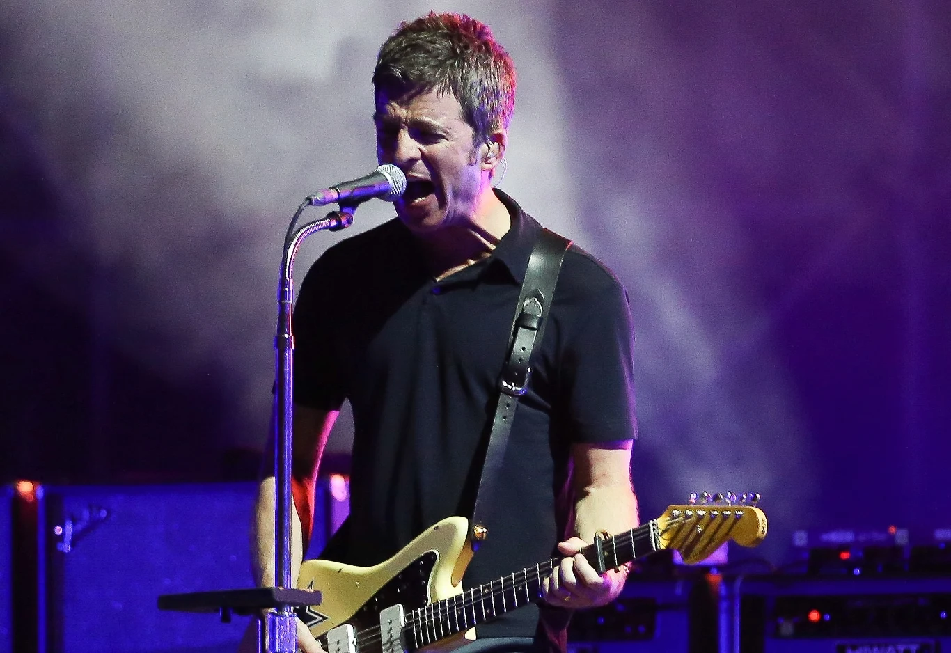 Noel Gallagher podczas koncertu Mężczyzna śpiewający do mikrofonu podczas występu na scenie, grający jednocześnie na gitarze elektrycznej, w tle widoczne światła sceniczne i sprzęt muzyczny.