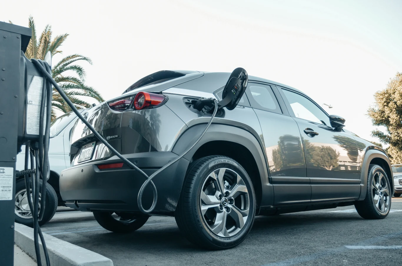 Program badawczy wart 14 mln dolarów potwierdził, że dane o zasięgu samochodów elektrycznych są zawyżone. Różnice sięgają od kilku do nawet 23 proc. w zależności od modelu. Elektryczny samochód typu SUV podłączony do stacji ładowania na zewnątrz, w pobliżu widoczne są palmy i inne zaparkowane pojazdy.