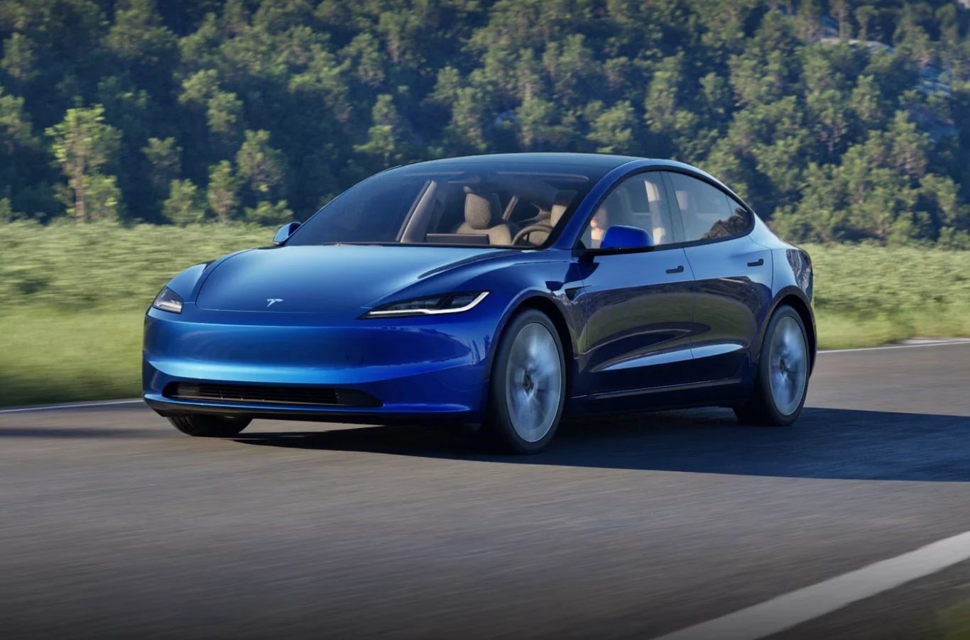 Tesla Model 3 przejechała 441 km zamiast deklarowanych 513 km. Niebieski samochód Tesla Model 3 poruszający się po drodze, otoczony zielonym krajobrazem z lasem w tle; samochód w dobrym stanie, nowoczesny wygląd, jasne wnętrze widoczne przez szyby.