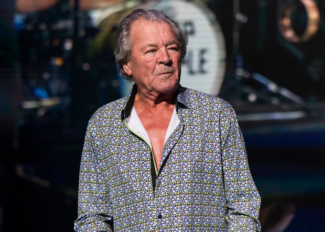 Ian Gillan jest wokalistą Deep Purple Mężczyzna w średnim wieku ubrany w koszulę z wzorem stoi na scenie przed rozmytym tłem z elementami perkusji.