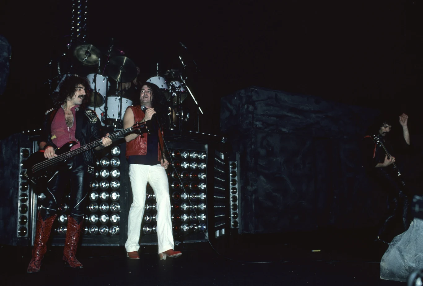 Ian Gillan w składzie Black Sabbath, z lewej Geezer Butler Zespół rockowy występujący na scenie, na pierwszym planie basista i wokalista, w tle widoczny perkusista na podwyższeniu, oświetlenie sceniczne skupia się na muzykach, otoczenie ciemne i industrialne, atmosfera dynamiczna i energetyczna.