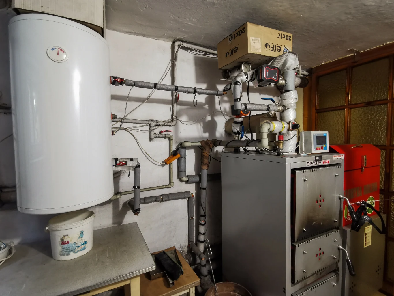 Z listy modeli kotłów, na które można dostać pieniądze z "Czystego Powietrza" zniknie 31 modeli Pomieszczenie techniczne z piecem i licznymi instalacjami hydraulicznymi, obok umieszczony biały bojler i różnorodne rury, na urządzeniach widoczne są przewody, czujniki i zawory.