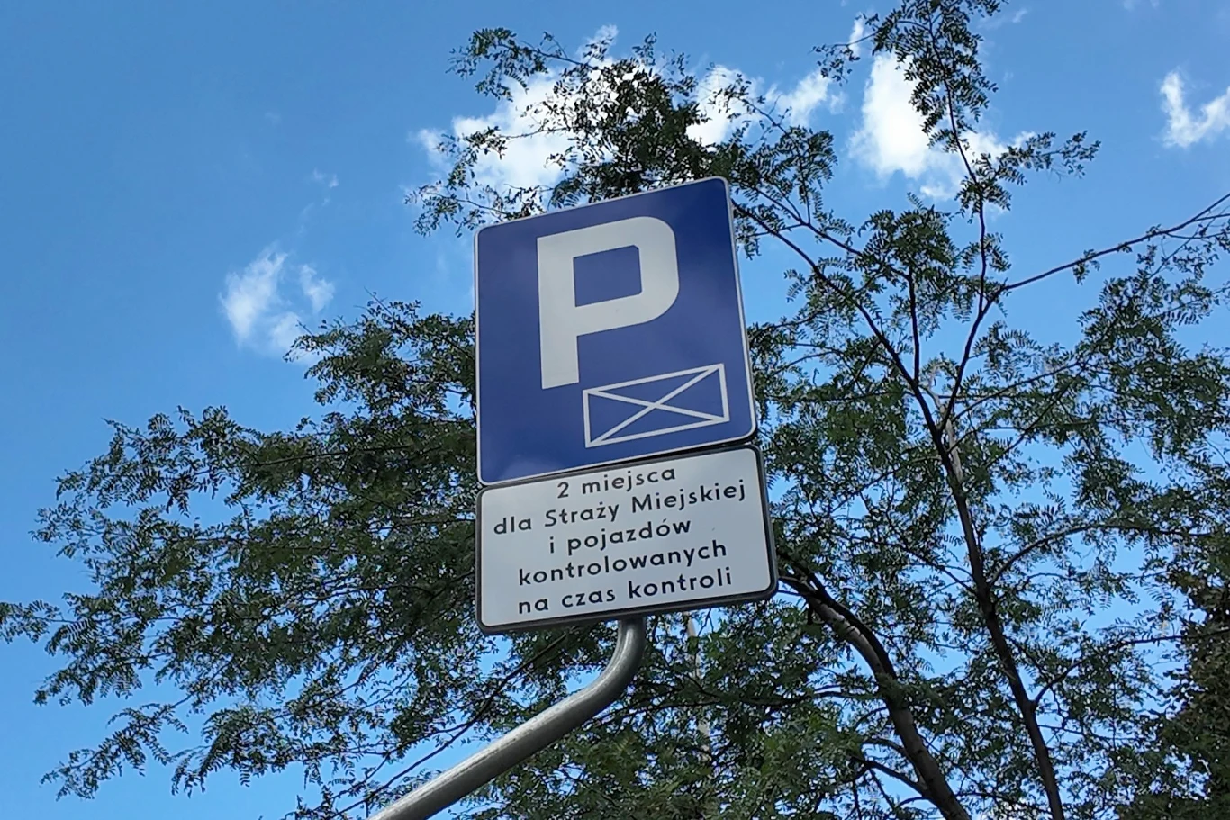 Znak drogowy informujący o wyznaczonym miejscu parkingowym dla Straży Miejskiej oraz pojazdów kontrolowanych na czas kontroli, z tłem nieba i konarami drzewa.
