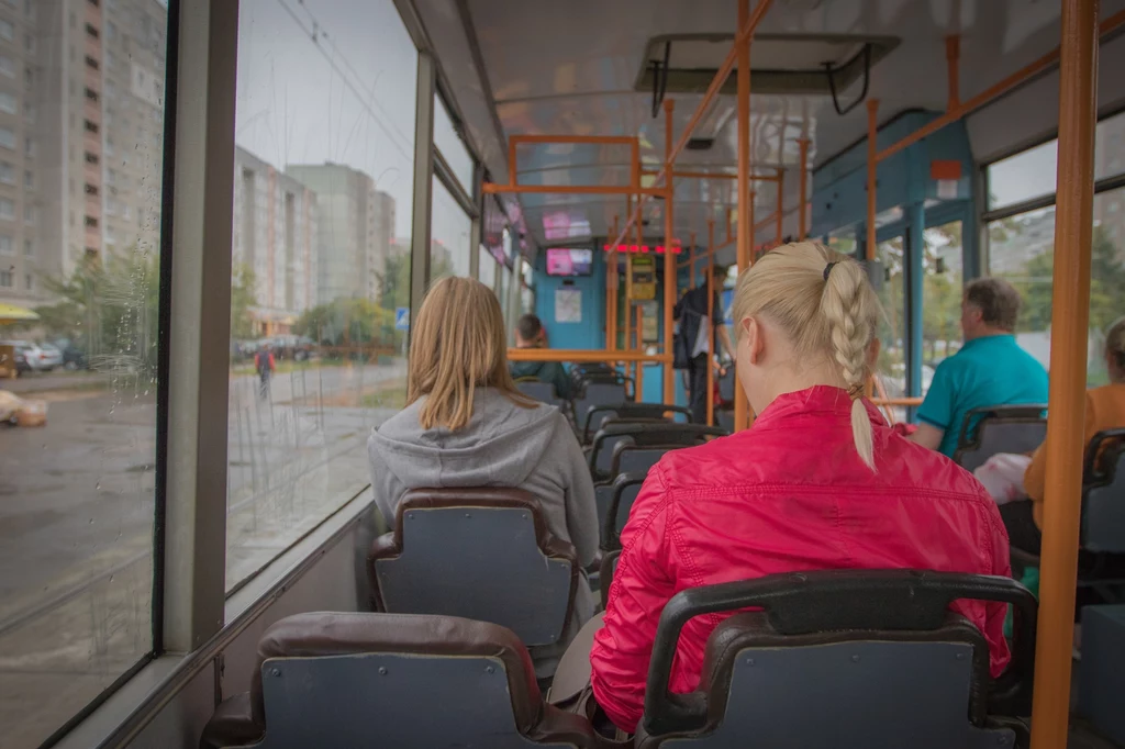 Wnętrze tramwaju z kilkoma pasażerami siedzącymi plecami do obiektywu, za oknem widać bloki mieszkalne oraz deszczową pogodę. Uwagę przyciąga kobieta w różowej kurtce z jasnym warkoczem.