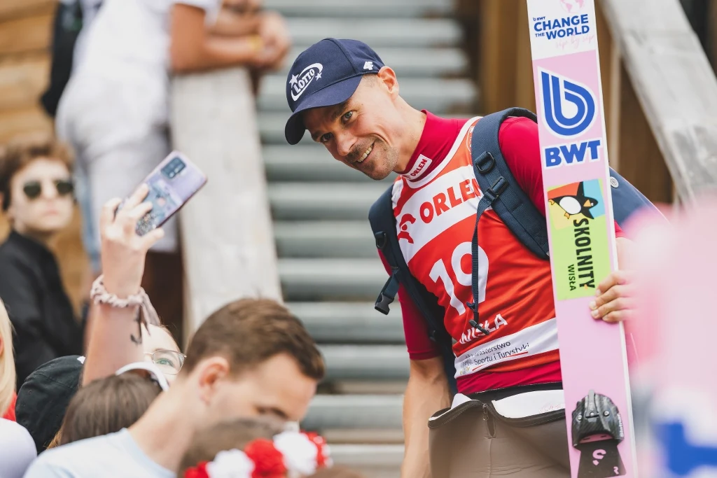 Zawodnik w sportowym stroju narciarskim trzymający narty, uśmiecha się do fanów, którzy robią z nim zdjęcia. W tle widoczna drewniana konstrukcja oraz kibice.