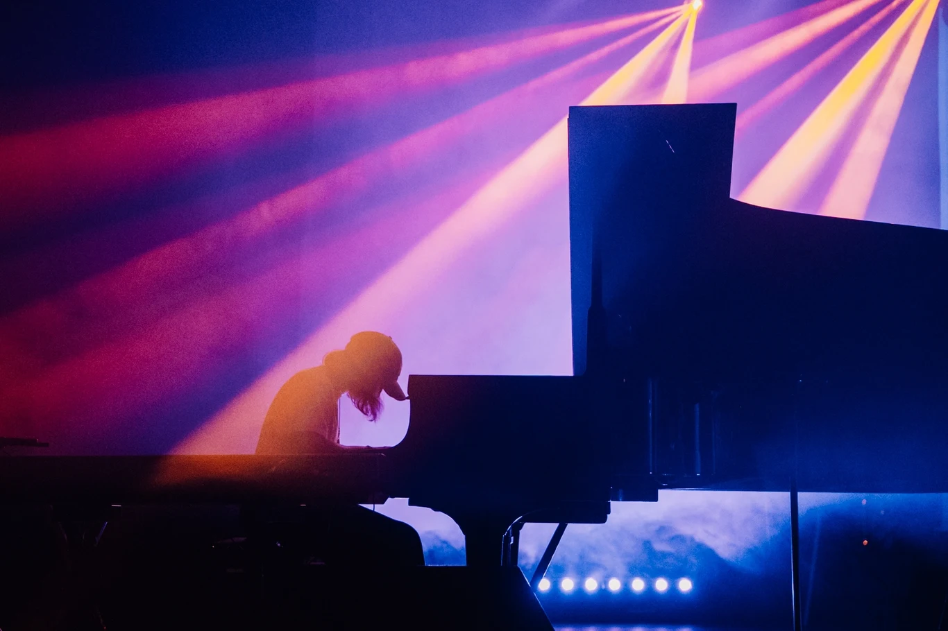 Sylwetka pianisty pochylonego nad fortepianem, oświetlona kolorowymi światłami scenicznymi w tle, dynamiczne promienie światła przecinają scenę, tworząc wyjątkowy klimat muzyczny.