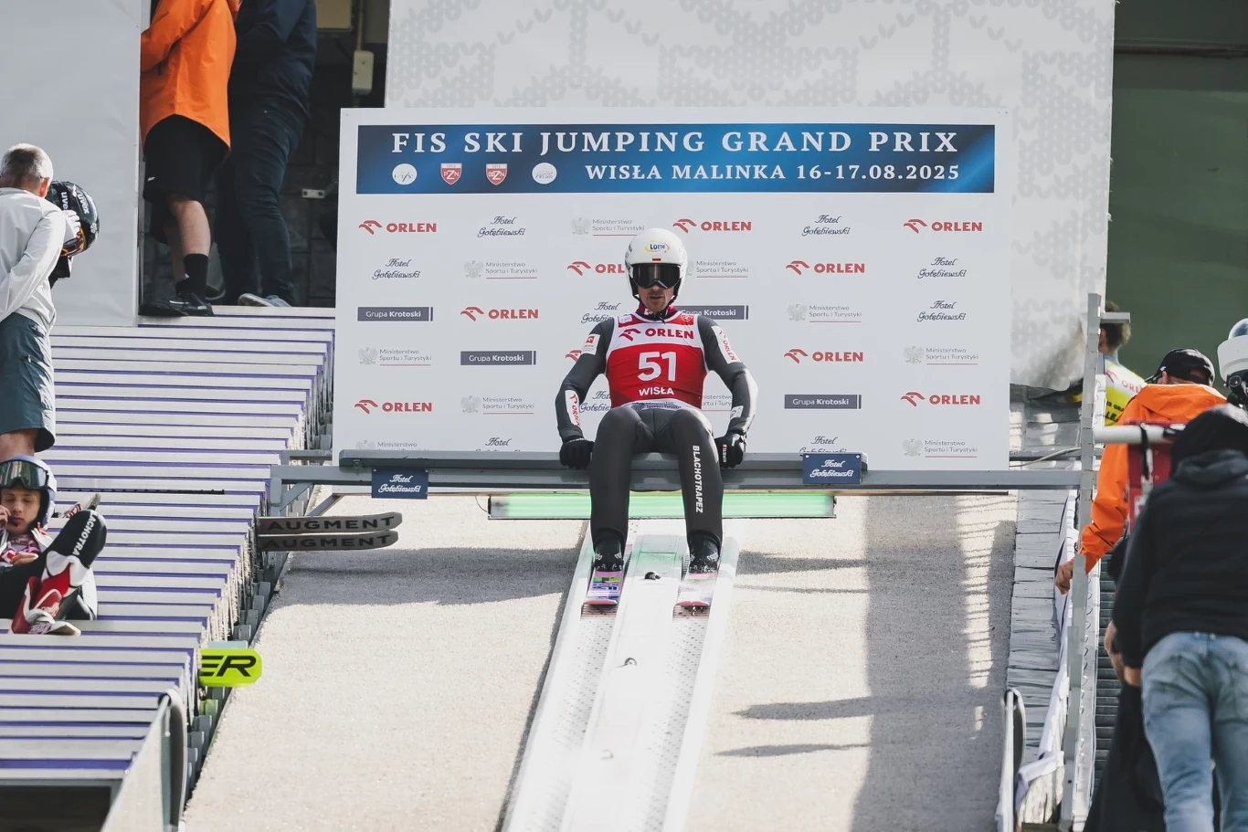 Zawodnik w stroju narciarskim z numerem 51 siedzi na belce startowej skoczni narciarskiej tuż przed startem, w tle widoczna tablica z informacjami o zawodach FIS Ski Jumping Grand Prix Wisła Malinka oraz osoby techniczne.
