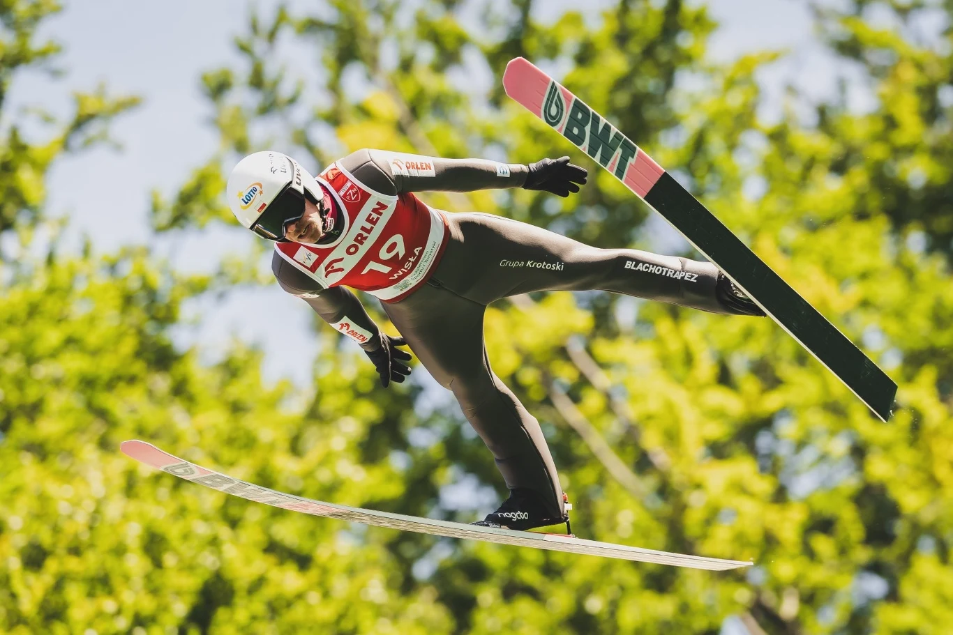 Skoczek narciarski w locie nad zielonym, rozmytym tłem drzewa, ubrany w kombinezon i kask, wyraźnie widoczne narty oraz numer startowy, dynamiczna pozycja w czasie skoku.