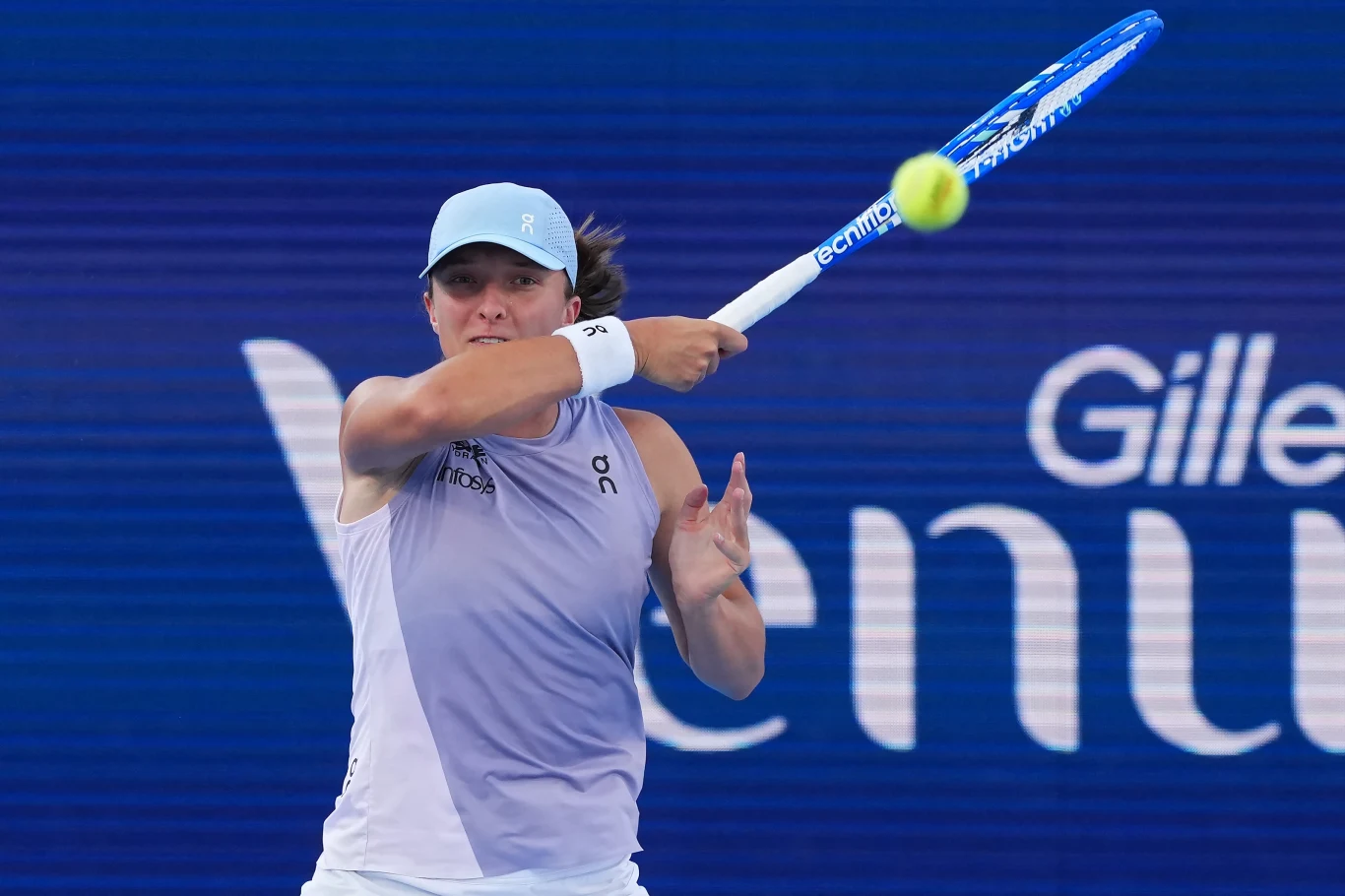 Iga Świątek Tenisistka wykonująca energiczny forhend, w tle widoczne logo sponsora i niebieskie boisko tenisowe.