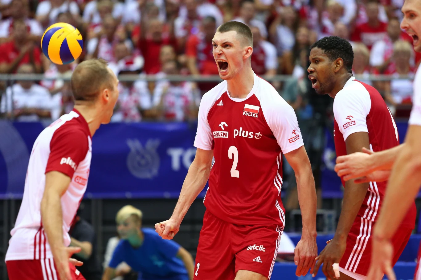 Emocjonująca scena podczas meczu siatkówki, trzej polscy siatkarze w czerwono-białych strojach celebrują zdobyty punkt, w tle widać rozemocjonowaną publiczność oraz lecącą piłkę ponad boiskiem.