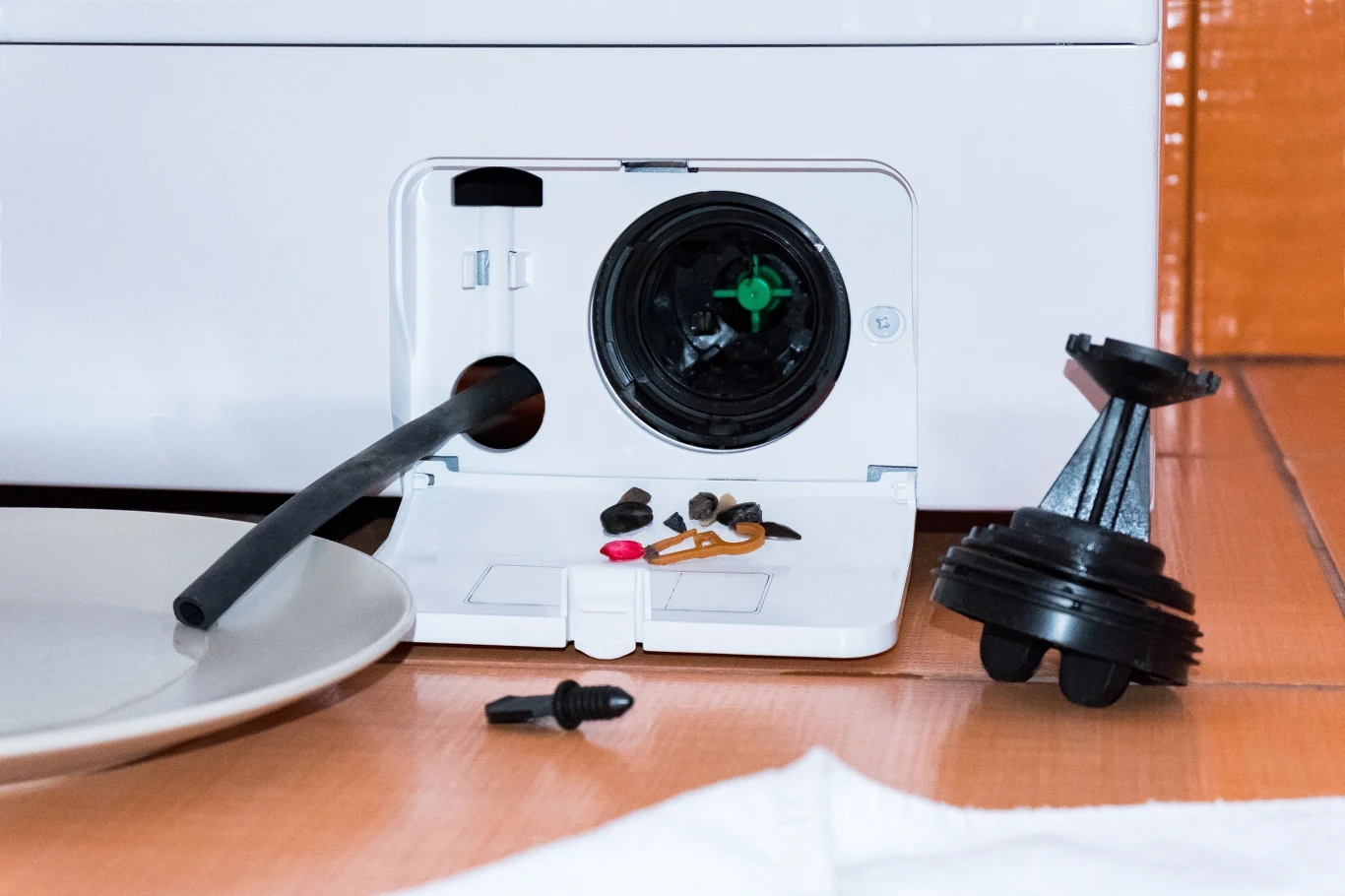 Otwarty filtr pralki z widocznymi śmieciami i drobnymi przedmiotami w komorze filtra, wyjęta pokrywa filtra i gumowa rurka do spuszczania wody na tle ceramicznej podłogi.