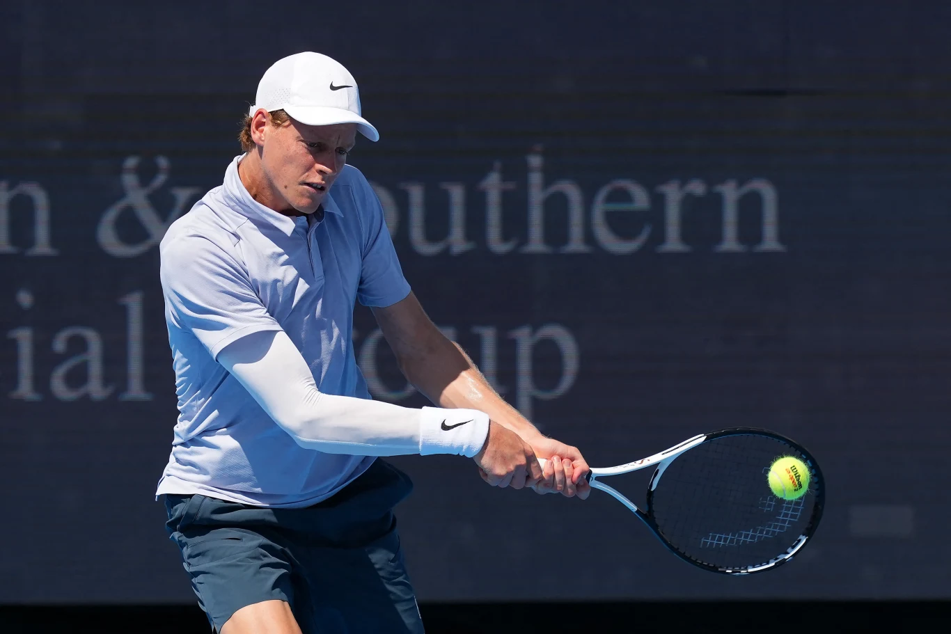 Tenisista ubrany w jasną koszulkę i czapkę, uderzający piłkę rakietą podczas meczu na korcie tenisowym, dynamiczna poza i skupienie na grze.