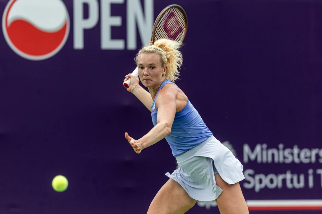 Katerina Siniakova Tenisistka w jasnym stroju sportowym uderza piłkę na korcie podczas meczu turniejowego, w tle widoczny baner z logo i napisem związanym z organizatorem zawodów.