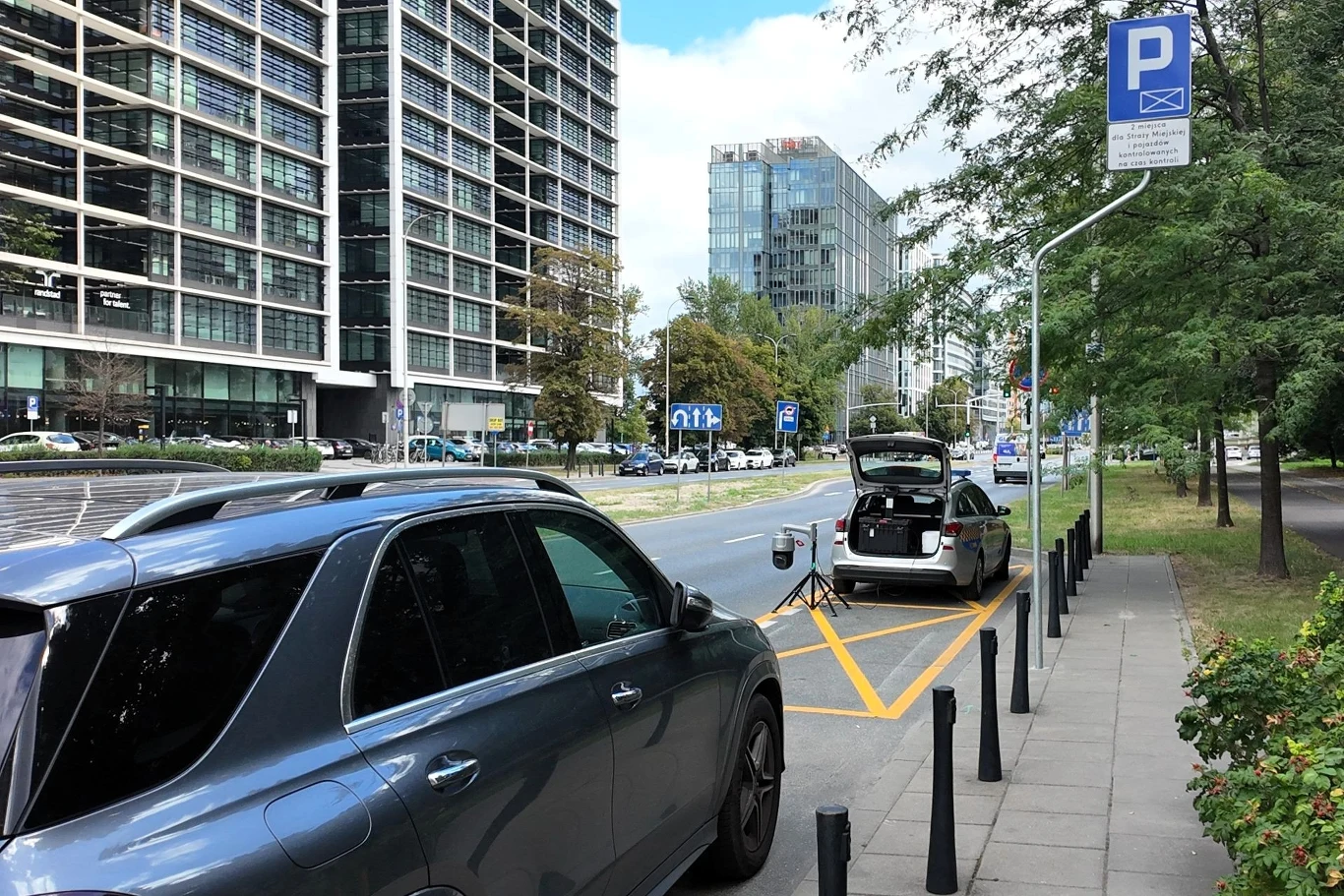 Czarna i srebrna nowoczesna osobówka zaparkowane przy ruchliwej ulicy w mieście, w tle wysokie budynki biurowe, znak z informacją o strefie płatnego parkowania, przy jednym z samochodów zamontowane urządzenie inspekcyjne.