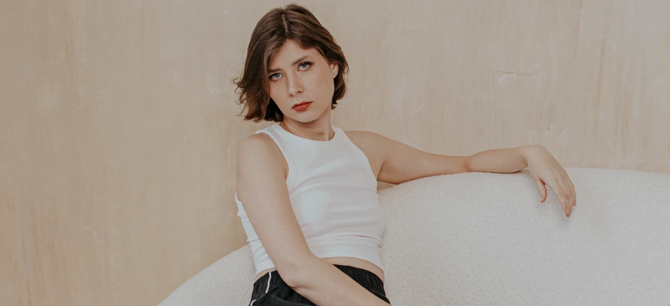 DOMI/NIKA Młoda kobieta z krótkimi brązowymi włosami siedzi na jasnej kanapie, ubrana w biały top i ciemne spodnie, patrzy bezpośrednio w obiektyw z poważnym wyrazem twarzy.