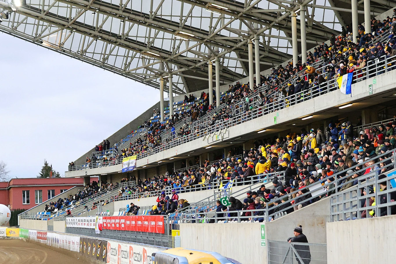 Tłum kibiców siedzących na trybunach stadionu, większość ubrana w klubowe barwy, niektóre osoby trzymają flagi, a w tle widoczna konstrukcja dachu oraz fragment budynku