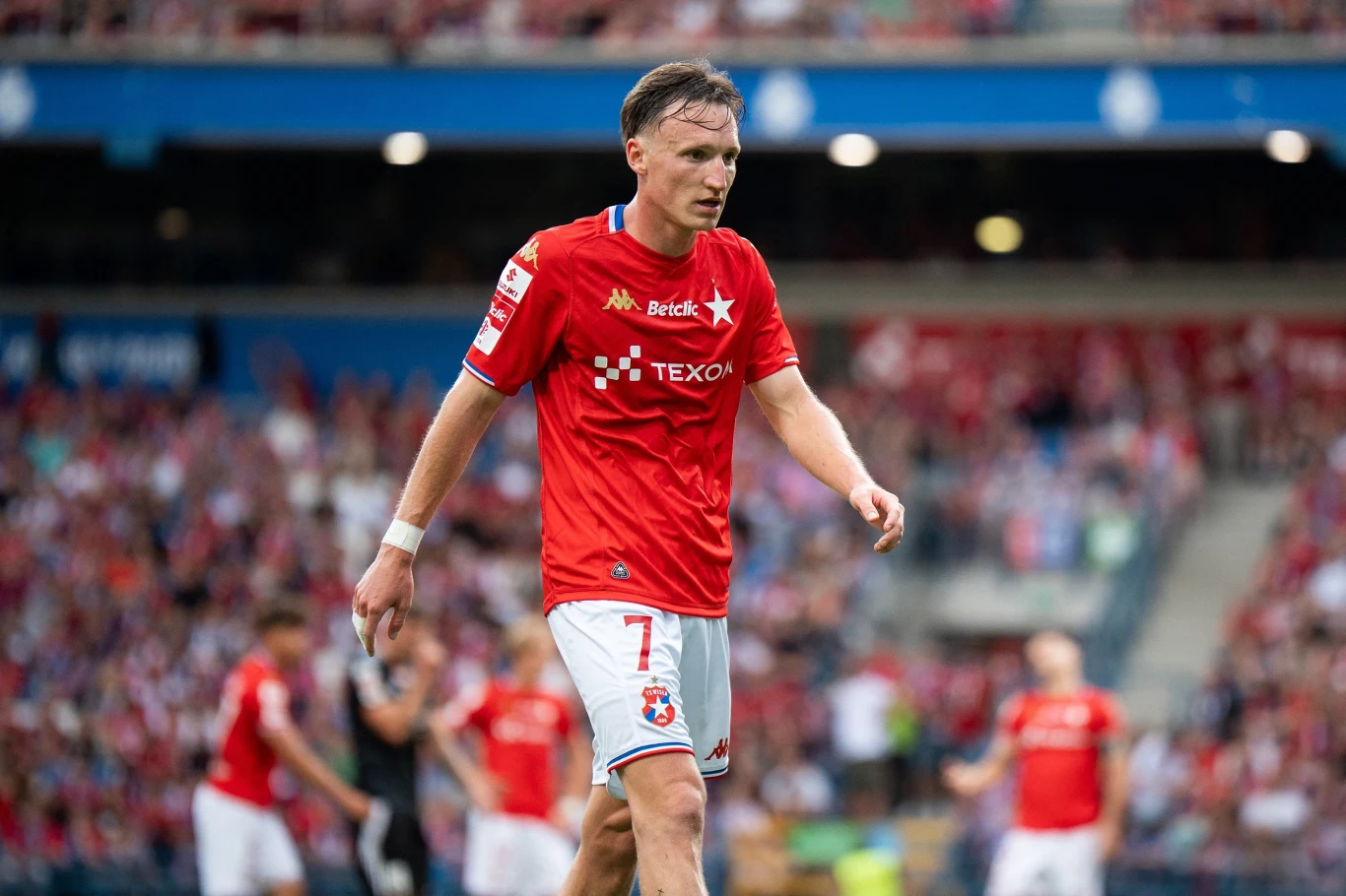 Piłkarz w czerwonej koszulce i białych spodenkach podczas meczu na stadionie, widoczni również inni zawodnicy i kibice na trybunach.