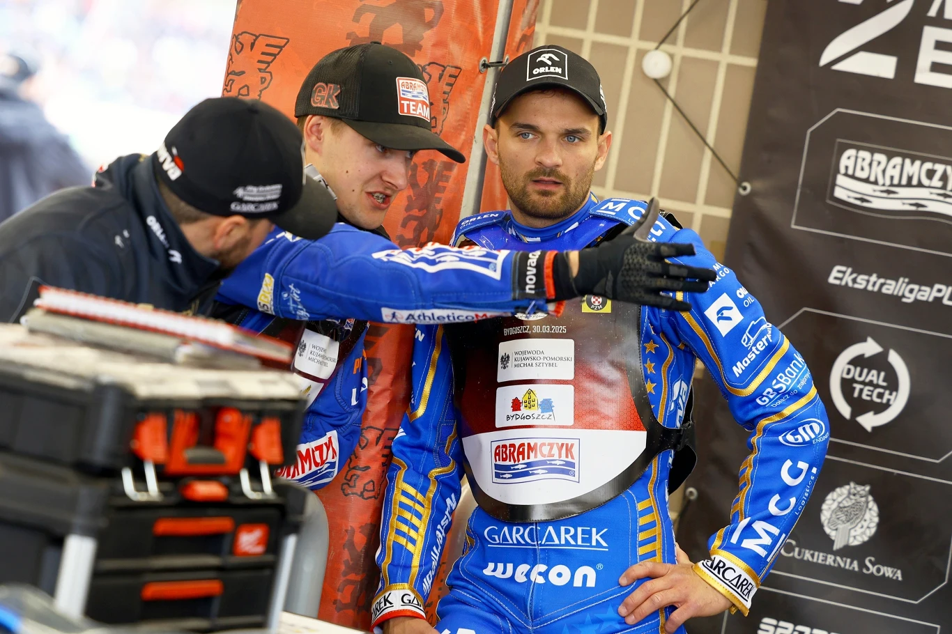 Trzech mężczyzn ubranych w sportowe stroje żużlowe w trakcie rozmowy, jeden z nich energicznie gestykuluje pokazując coś poza kadrem, w tle widoczne banery z logo sponsorów oraz skrzynka narzędziowa.