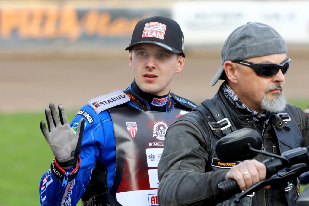 Wiktor Przyjemski Zawodnik żużlowy w stroju z licznymi logotypami sponsorskimi stoi obok motocykla, podnosząc rękę w geście powitania. Obok niego mężczyzna w czarnych okularach i czapce prowadzi motocykl.