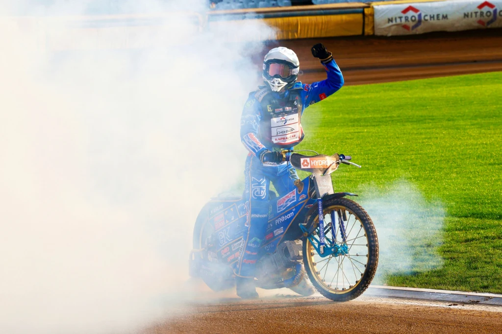 Wiktor Przyjemski, cieszący się ze złota MIMP 2025 Zawodnik żużlowy w niebieskim kombinezonie i kasku unosi rękę w geście pozdrowienia, stojąc na motorze otoczonym gęstym dymem na torze żużlowym, w tle widać zieloną murawę oraz fragment trybun.