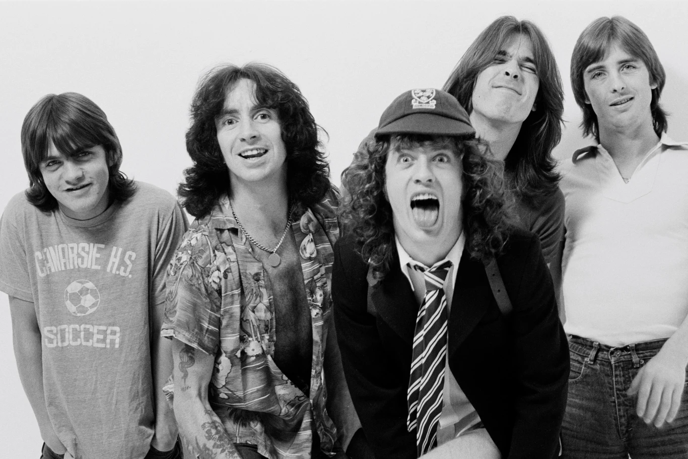 Malcolm Young, Bon Scott, Angus Young, Cliff Williams i Phil Rudd z AC/DC Pięciu mężczyzn stojących razem, uśmiechają się i pokazują luźne, wesołe postawy, jeden z nich wyróżnia się charakterystyczną czapką i szeroko otwartymi ustami, reszta ubrana jest w casualowe koszulki.