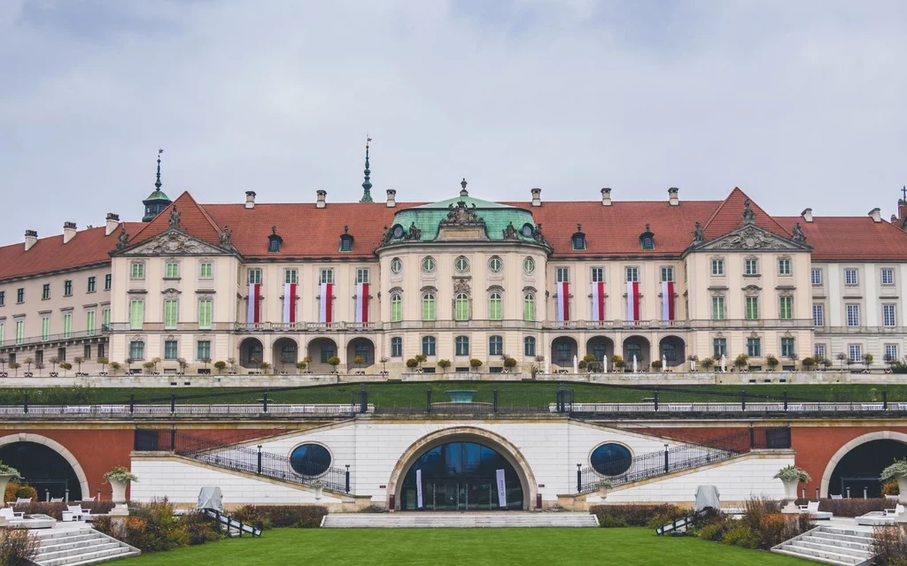Zamek Królewski w Warszawie Zamek Królewski w Warszawie z czerwoną dachówką, zieloną kopułą pośrodku oraz białą fasadą ozdobioną licznymi flagami Polski i symetrycznymi ogrodami na pierwszym planie.