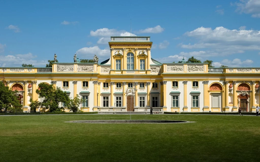 Pałac w Wilanowie Barokowy pałac z żółto-białą fasadą, bogato zdobiony detalami architektonicznymi, otoczony rozległym trawnikiem i niewielką fontanną pośrodku dziedzińca, w pogodny dzień z niebieskim niebem i białymi chmurami.