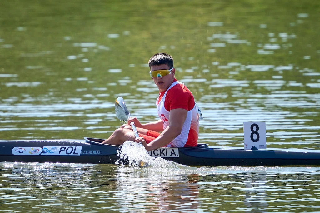 Kajakarz w biało-czerwonym stroju sportowym płynie jedynką po spokojnej wodzie, wykonując dynamiczny ruch wiosłem, na łodzi widoczny numer startowy 8 oraz nazwisko zawodnika i oznaczenie kraju Polska.