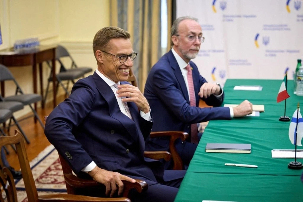 Dwóch mężczyzn w garniturach siedzi przy zielonym stole konferencyjnym, na którym stoją niewielkie flagi państwowe, notesy i butelki wody, w tle baner konferencyjny oraz eleganckie wnętrze sali spotkań.