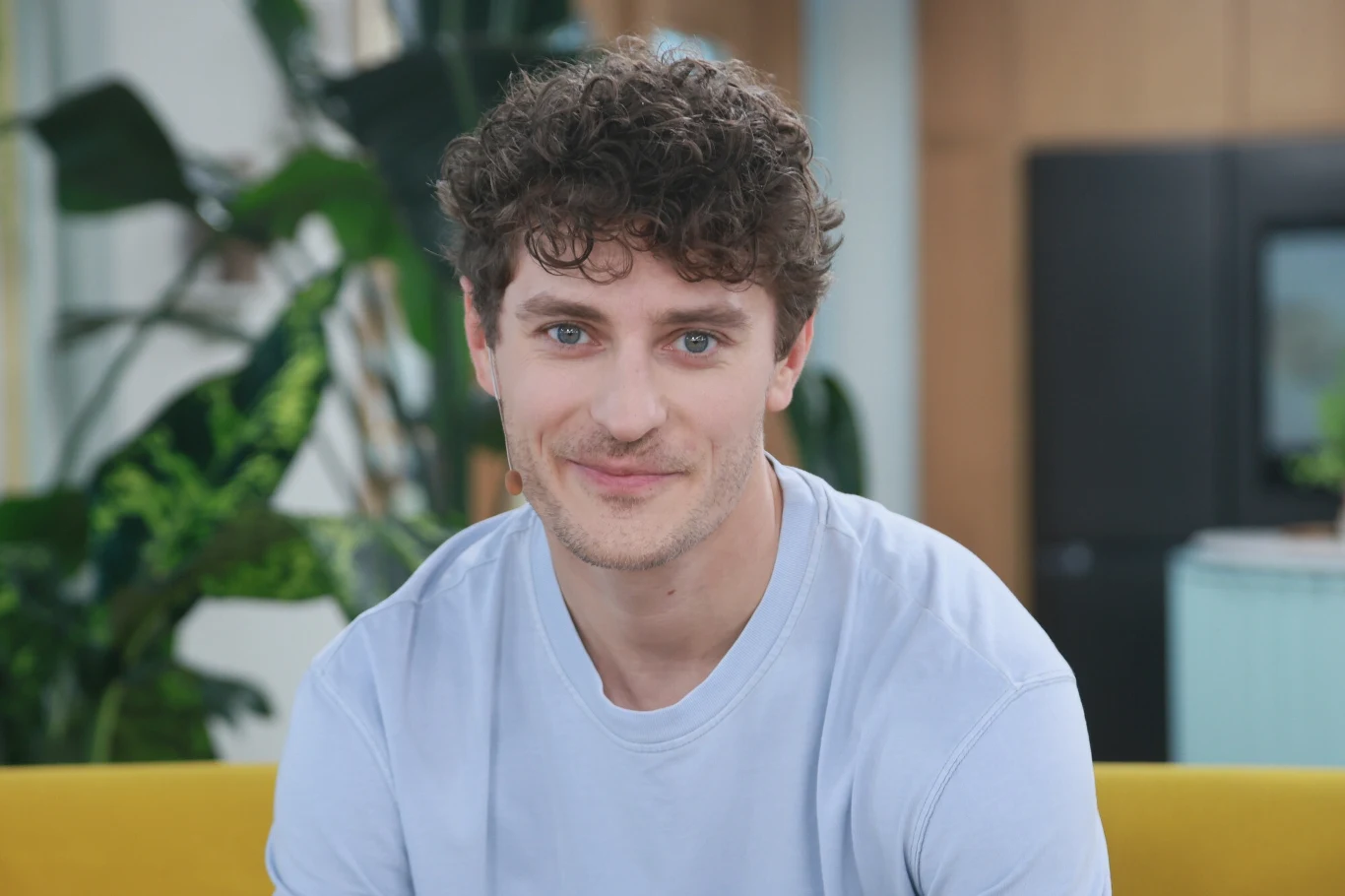 Młody mężczyzna z kręconymi włosami uśmiecha się delikatnie, ubrany w jasnoniebieski t-shirt, w tle widoczne są zielone rośliny i nowoczesne wnętrze mieszkania lub biura.