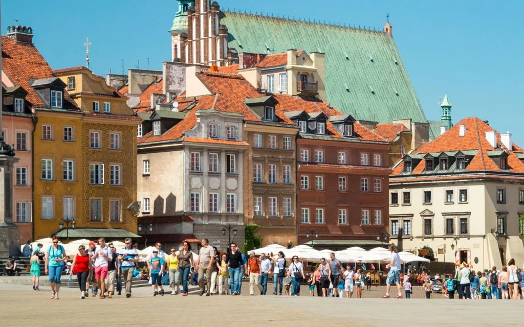 Warszawa każdego roku przyjmuje coraz to więcej turystów z Polski i z zagranicy Kolorowe kamienice Starego Miasta w Warszawie, tętniący życiem plac z licznymi turystami i mieszkańcami, tło stanowi dach katedry oraz niebieskie niebo.