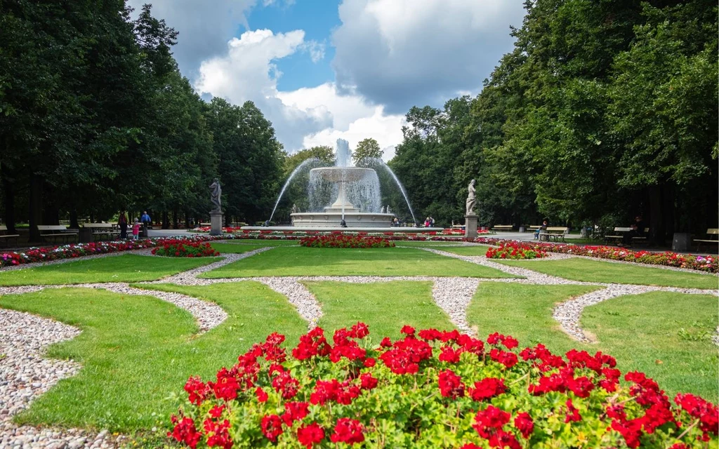 Ogród Saski Fontanna w centrum francuskiego ogrodu otoczona czerwonymi kwiatami i zadbanym trawnikiem, w tle aleje wysadzane drzewami, nad całością błękitne niebo z białymi chmurami.