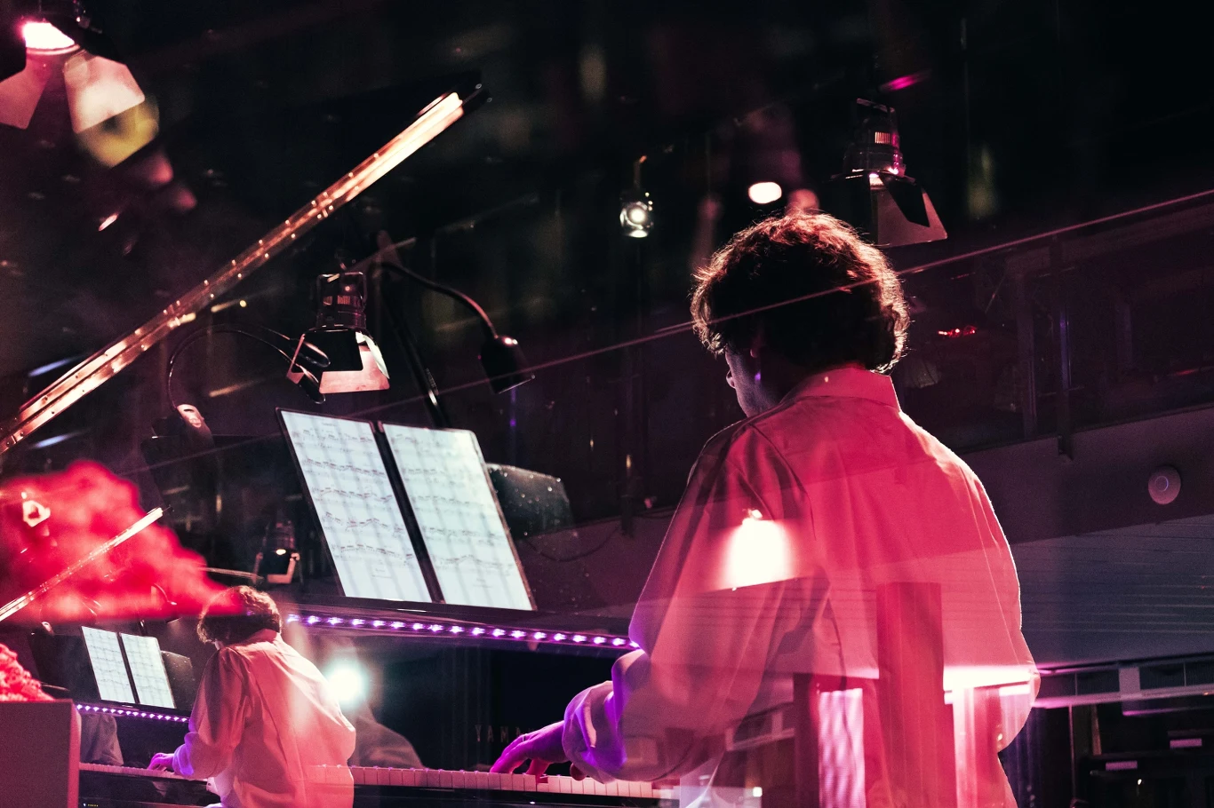 Muzyk grający na keyboardzie w sali koncertowej, ściany odbijają światło, a w tle widoczne są pulpity z nutami oraz sceniczne oświetlenie.