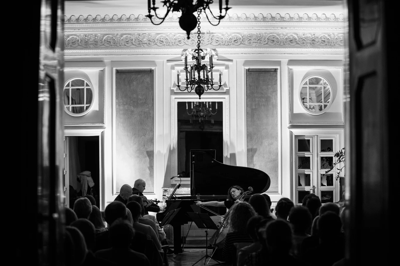 Koncert fortepianowy odbywający się w eleganckiej sali z klasycznymi zdobieniami, widownia skupiona na muzykach grających na fortepianie, z ozdobnym żyrandolem i światłem podkreślającym atmosferę wieczoru.