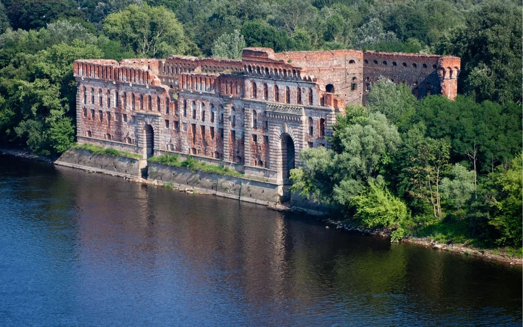 Twierdza Modlin Stary ceglany fort położony nad wodą, otoczony gęstą zielenią drzew. Budynek znajduje się tuż przy linii brzegu i sprawia wrażenie opuszczonego, z widocznymi licznymi oknami i elementami architektonicznymi typowymi dla fortyfikacji.