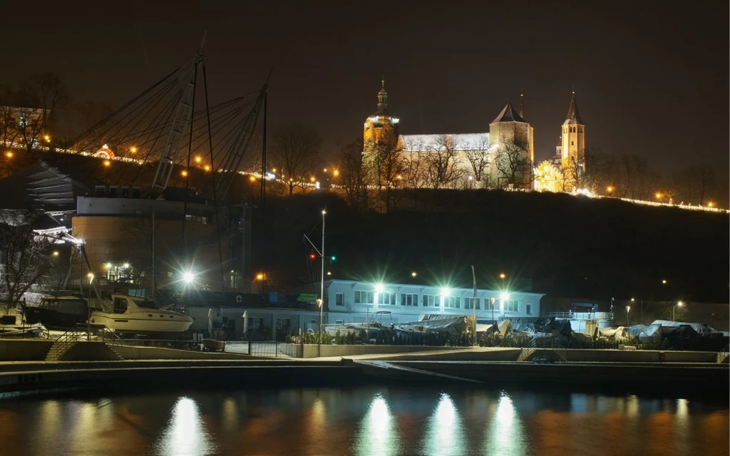 Płock nocą Zabytkowa katedra na wzgórzu nad rzeką, oświetlona nocą, w tle widoczne drzewa oraz łodzie i budynki portowe na pierwszym planie.