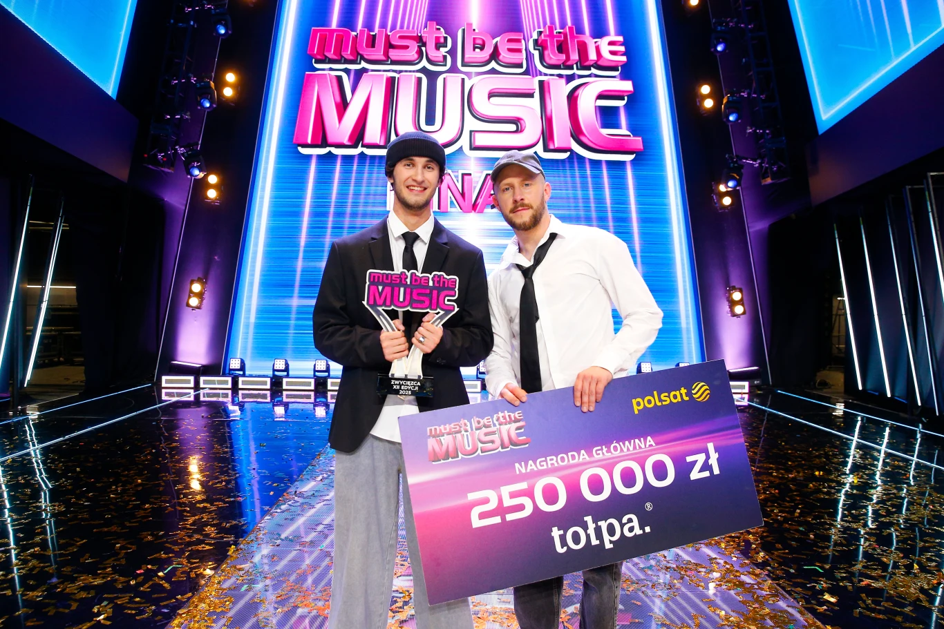 Alien i Majtis wygrali ostatnią edycję "Must Be The Music" Dwaj mężczyźni trzymają wielki czek na 250 000 zł oraz statuetkę programu Must Be The Music stojąc na scenie finałowej popularnego telewizyjnego show muzycznego, otoczeni światłami i neonami z logo programu.