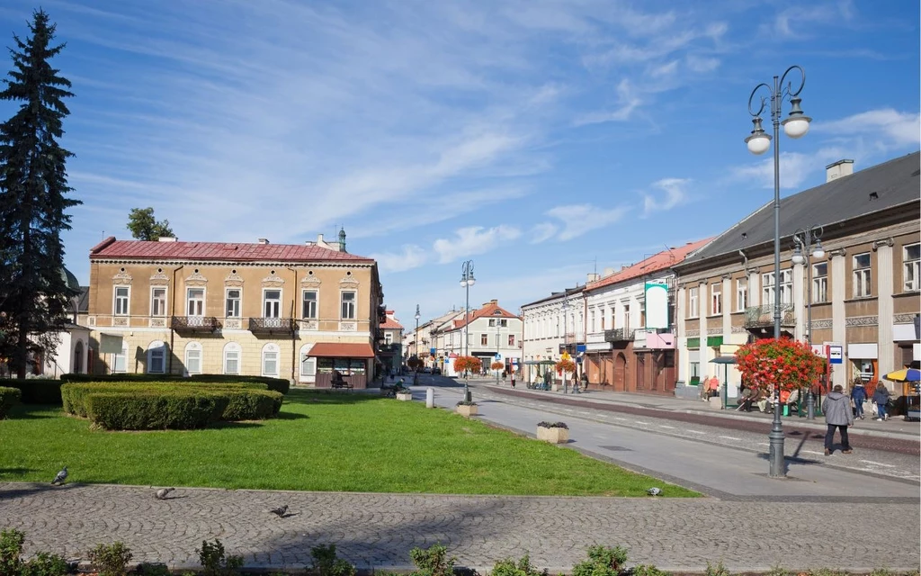 Radom Historyczna zabudowa rynku miejskiego z zadbanym trawnikiem i rabatą kwiatową, otoczonym kamienicami o jasnych elewacjach, brukowanym chodnikiem i dekoracyjnymi latarniami, na tle niebieskiego nieba.