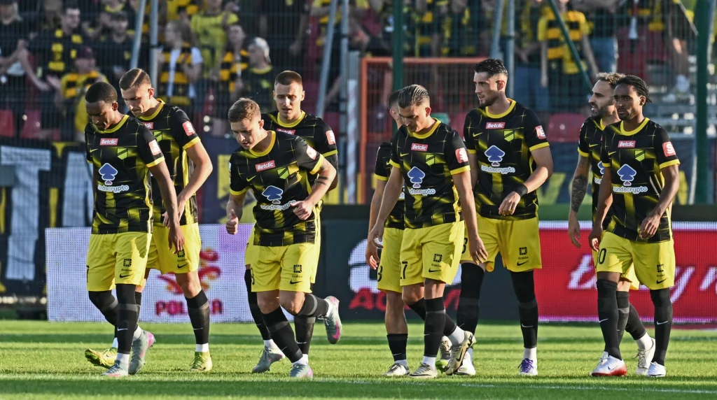 Piłkarze Wieczystej Kraków Grupa piłkarzy w żółto-czarnych strojach drużyny idąca razem po murawie boiska, w tle trybuny z kibicami ubranymi w podobnych barwach oraz ogrodzenie stadionu.