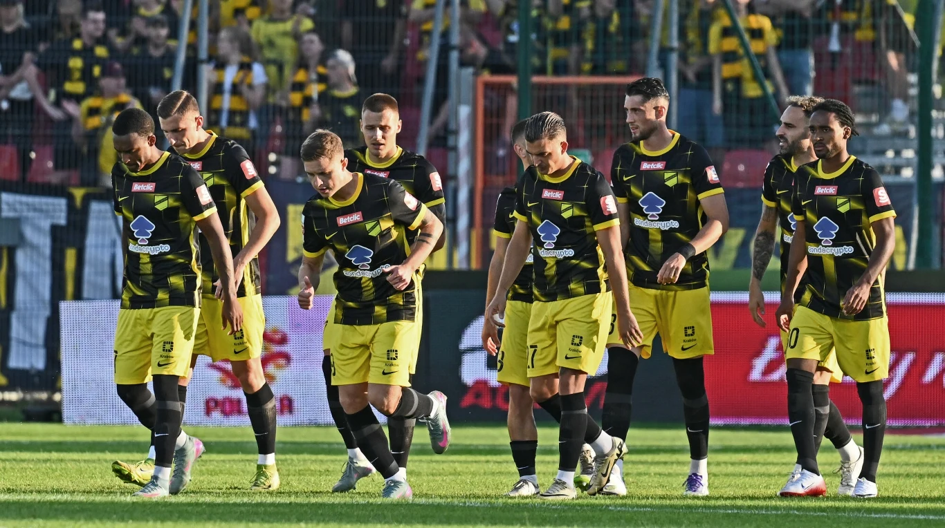 Grupa piłkarzy w żółto-czarnych strojach drużyny idąca razem po murawie boiska, w tle trybuny z kibicami ubranymi w podobnych barwach oraz ogrodzenie stadionu.