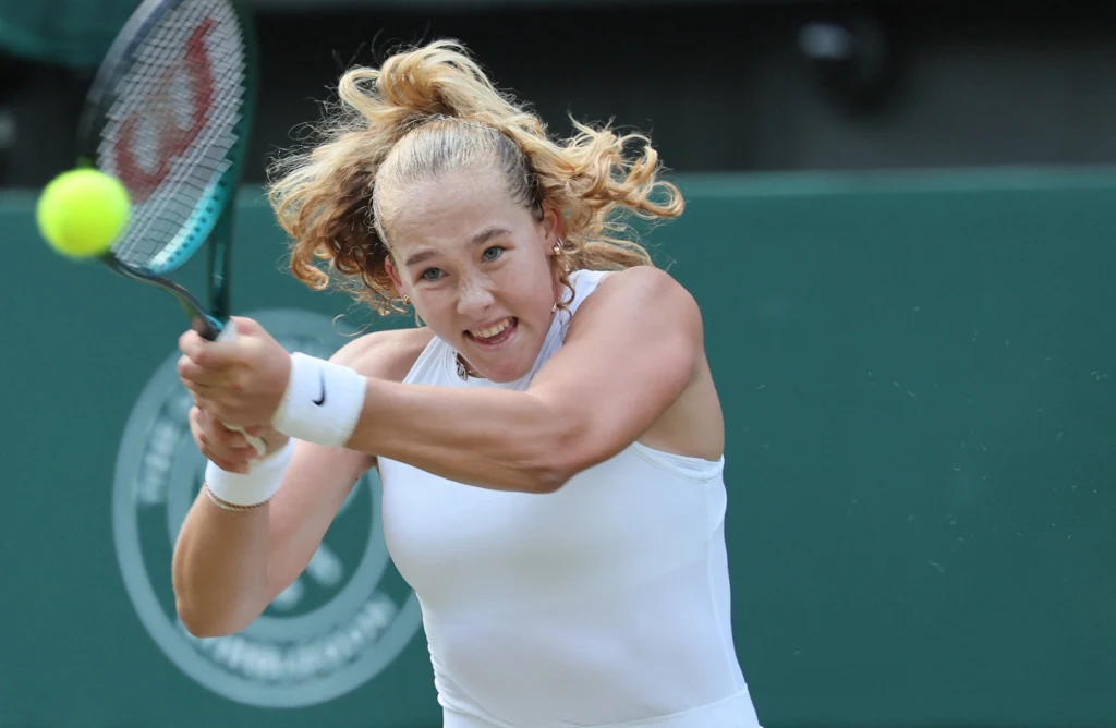 Mirra Andriejewa Tenisistka w białym stroju skupiona na grze, wykonuje energiczne uderzenie dwuręcznym backhandem, żółta piłka tenisowa zbliża się do rakiety, na tle zielonego kortu widoczne logo turnieju.