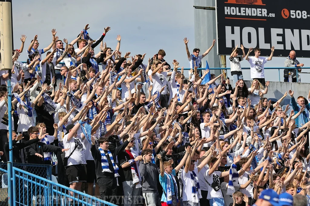 Kibice Unii Leszno Tłum kibiców ubranych w klubowe barwy zebrał się na trybunach stadionu, entuzjastycznie unosząc ręce do góry podczas wspólnego dopingu, wokół widoczna jest energia i pozytywna atmosfera sportowego wydarzenia.