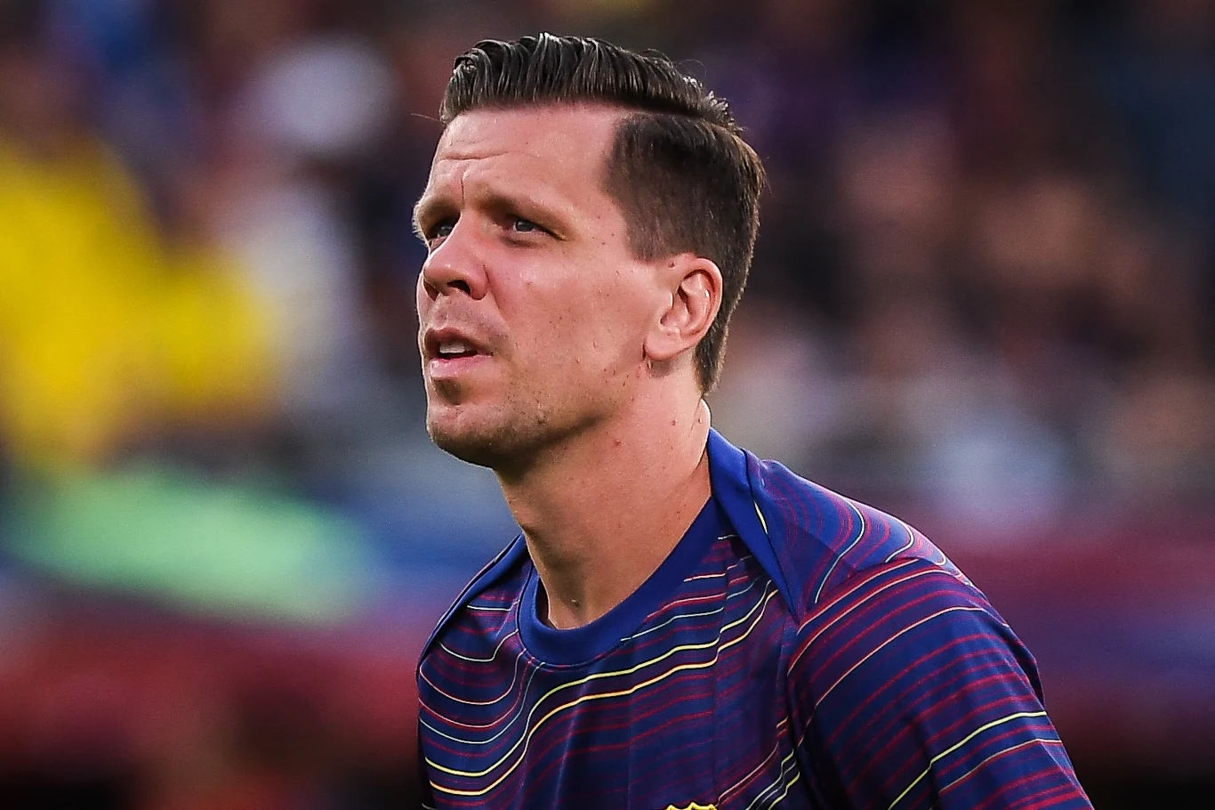 Wojciech Szczęsny w FC Barcelonie Piłkarz ubrany w koszulkę sportową z barwnymi wzorami stoi na tle rozmytego stadionu, skupiony spoglądając lekko w bok.