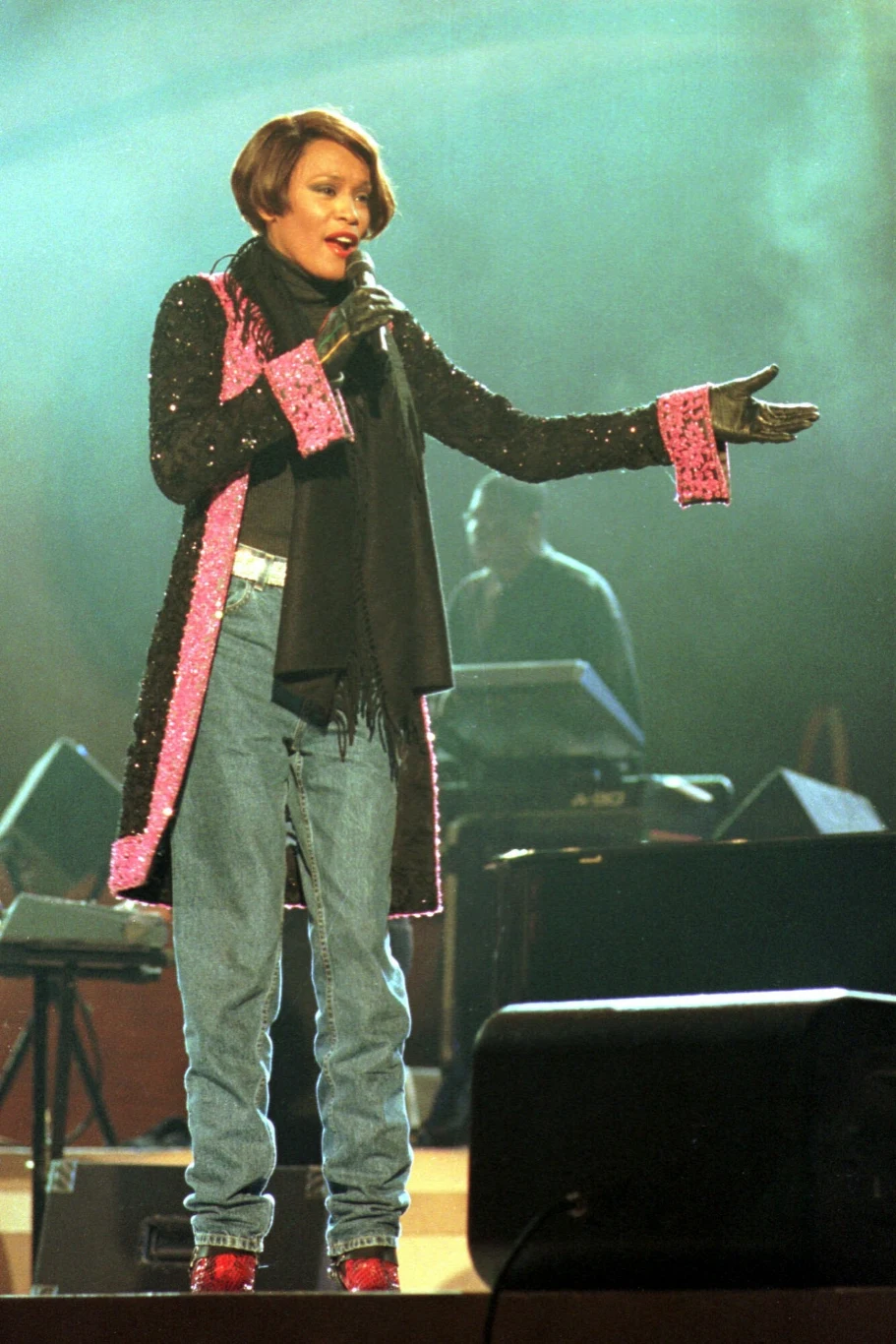 Kobieta ubrana w świecącą, cekinową marynarkę, czarny szalik oraz jeansy stoi na scenie, śpiewając do mikrofonu i gestykulując jedną ręką, z tyłu widoczny jest zamazany muzyk przy sprzęcie dźwiękowym.